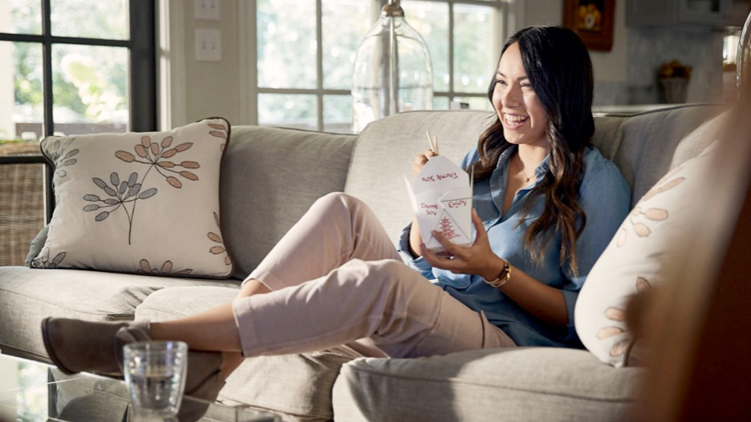 A woman sits on a sofa, smiling as she eats takeout with chopsticks. The room is bright with sunlight, and decorative pillows feature leaf patterns.