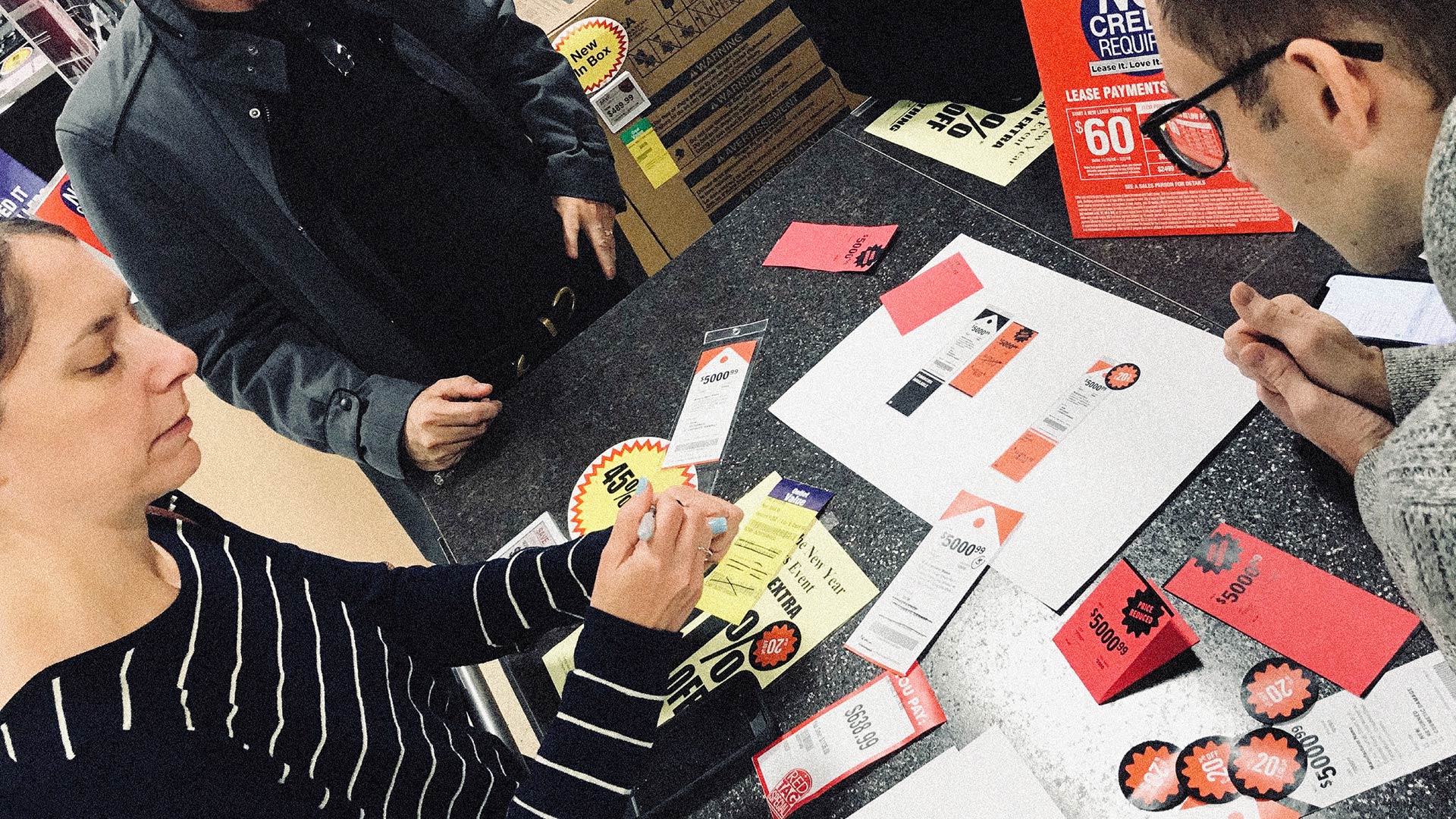 Two people collaborate at a desk covered with colorful flyers, papers, and circular tags. The mood is focused and collaborative, suggesting a creative project.