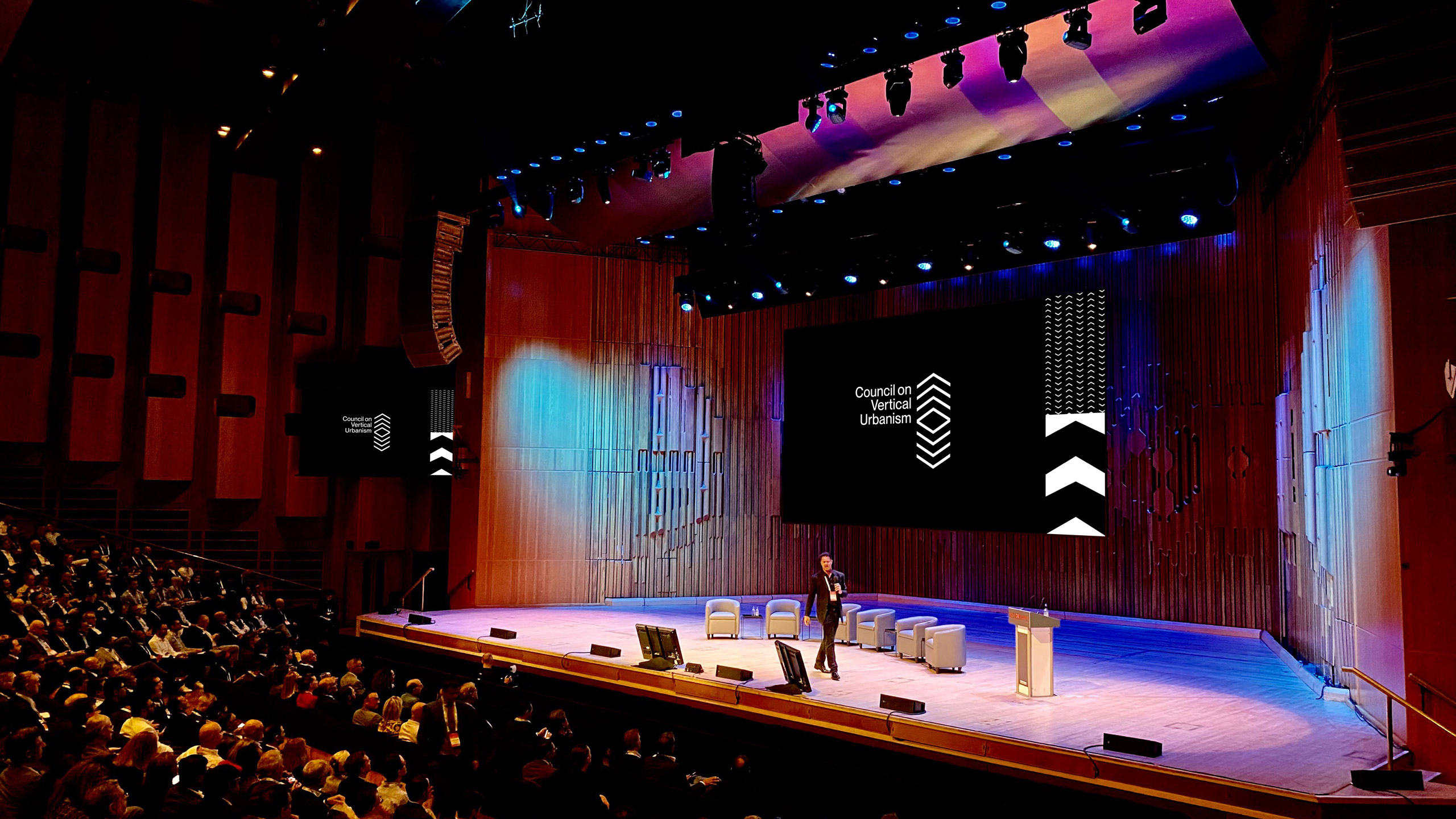 A speaker presents on stage to a large audience. A screen behind him displays "Council on Vertical Urbanism."